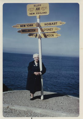 Photograph of Jean Milligan in New Zealand beside a sign-post pointing to New York, Wellington, H...